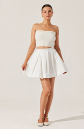 A woman is wearing a strapless, cream-colored, textured crop top with a banded elastic top edge. She pairs it with a high-waisted, white, pleated skirt that has a wide waistband and features box pleats for added volume. She completes the outfit with cream-colored closed-toe heels.